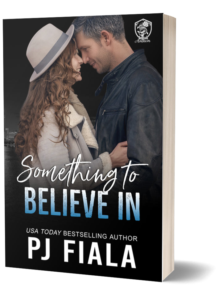  Paperback edition of Something to Believe In by PJ Fiala standing upright. The cover features a curly-haired woman wearing a light-colored hat and coat embracing a silver-haired man in a leather jacket against a nighttime city skyline. Part of the Blossom Springs: Rogue Anthem series.